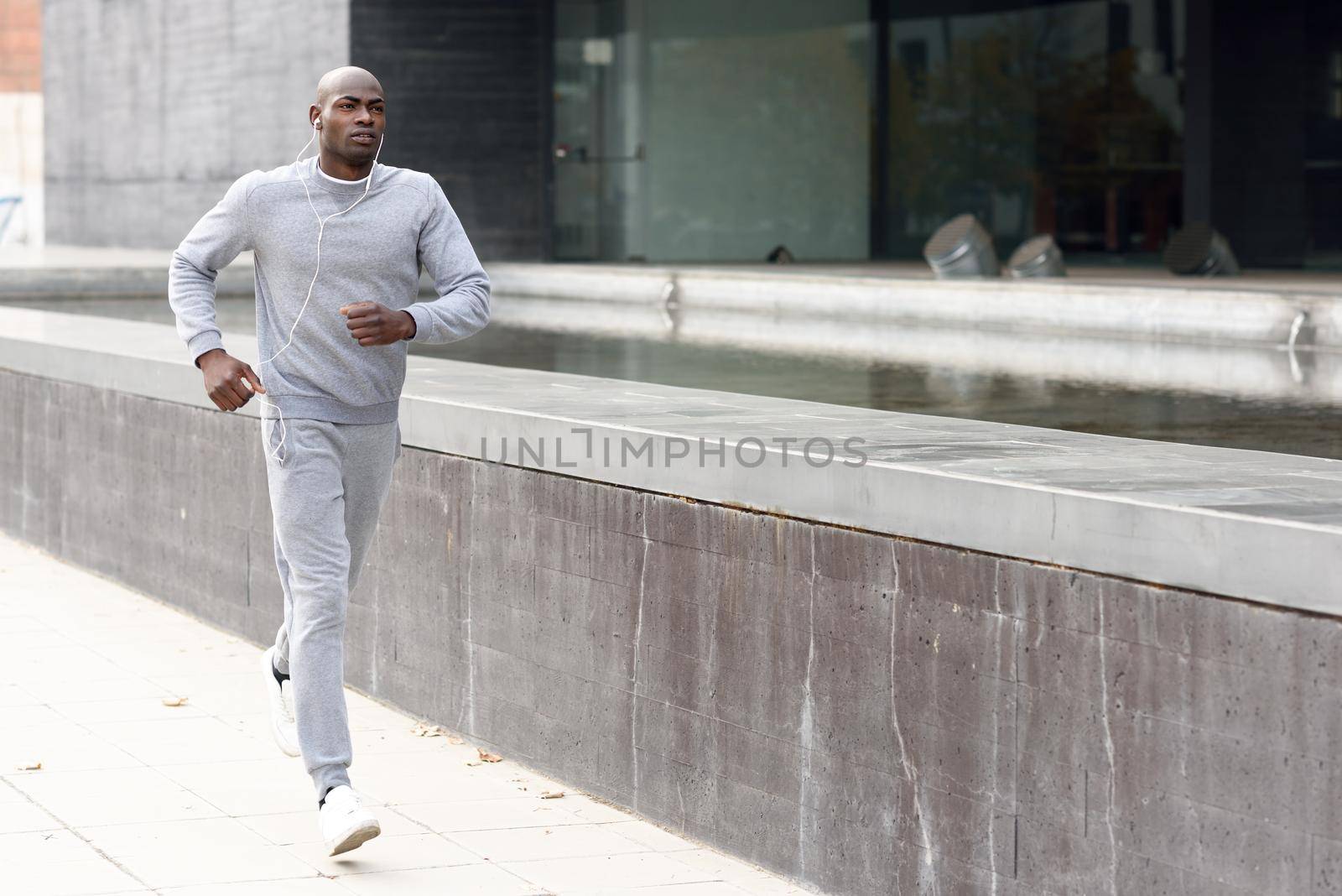 Attractive black man running in urban background. Male doing workout outdoors. Guy listening to music with white headphones
