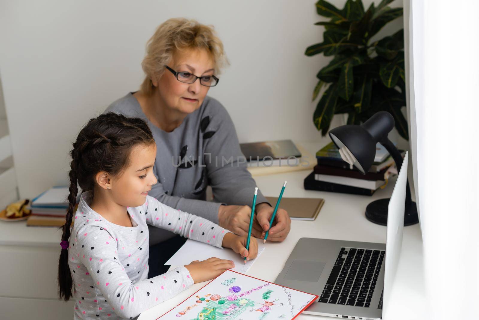 Mature grandmother helping child with homework at home. Satisfied old grandma helping her granddaughter studying in living room. Little girl writing on notebook with senior teacher sitting next to her by Andelov13