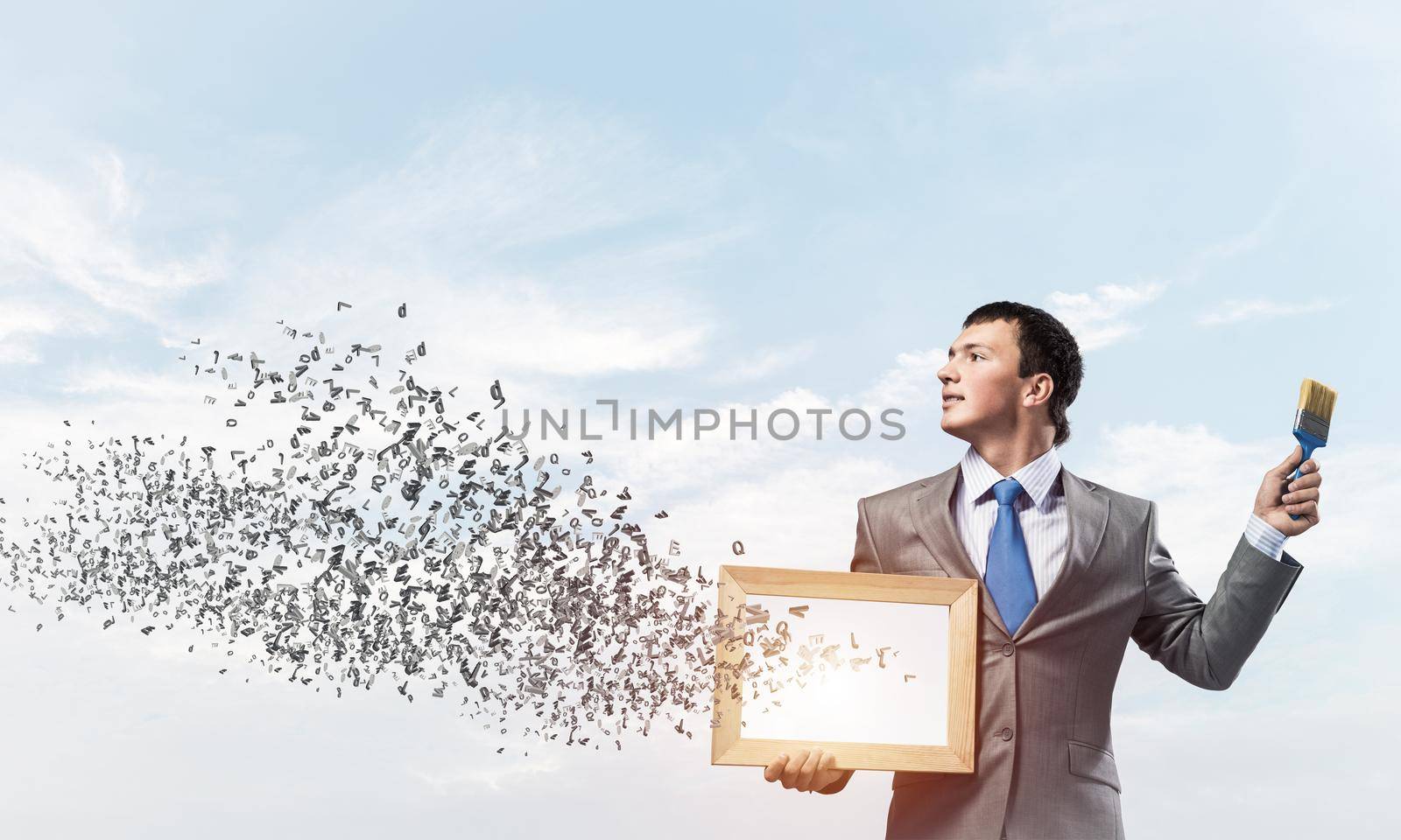 Creative businessman painter holding paint brush and whiteboard in hands. Portrait of happy handsome man in business suit and tie on blue sky background. Various letters flying out from canvas.