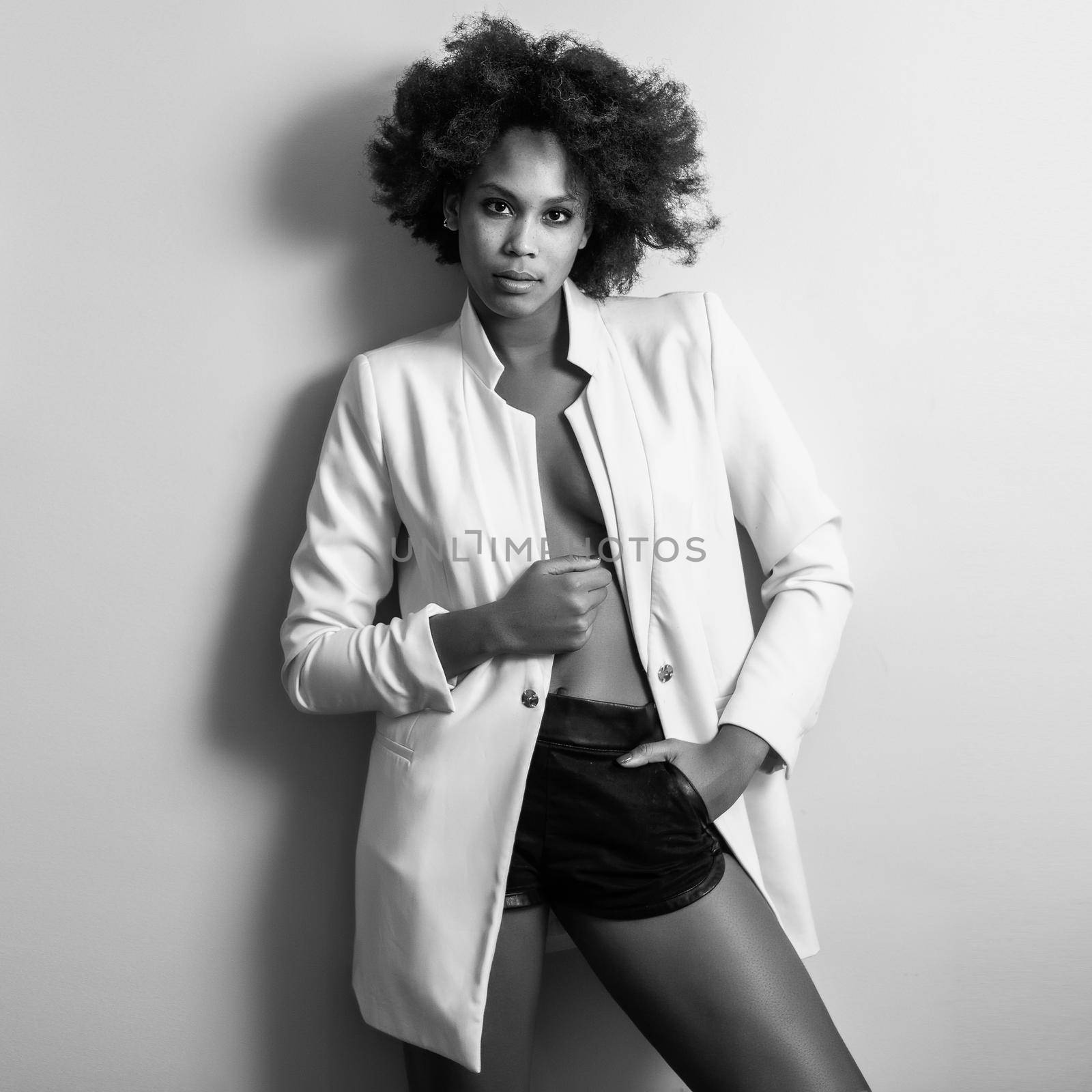 Young black woman with afro hairstyle possing near a white wall in her room. Mixed girl wearing blazer jacket and shorts.