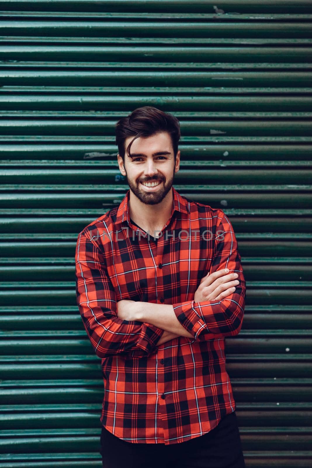 Young bearded man, model of fashion, wearing a plaid shirt with a green blind behind him. Smiling guy with beard and modern hairstyle in urban background.