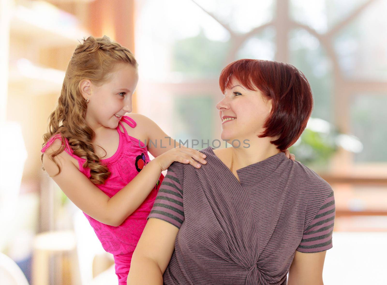 Happy family of two people,an adult mother, her beloved daughter 7 years. Daughter gently hugs the mother's neck. Close-up.In a room with a large semi-circular window.