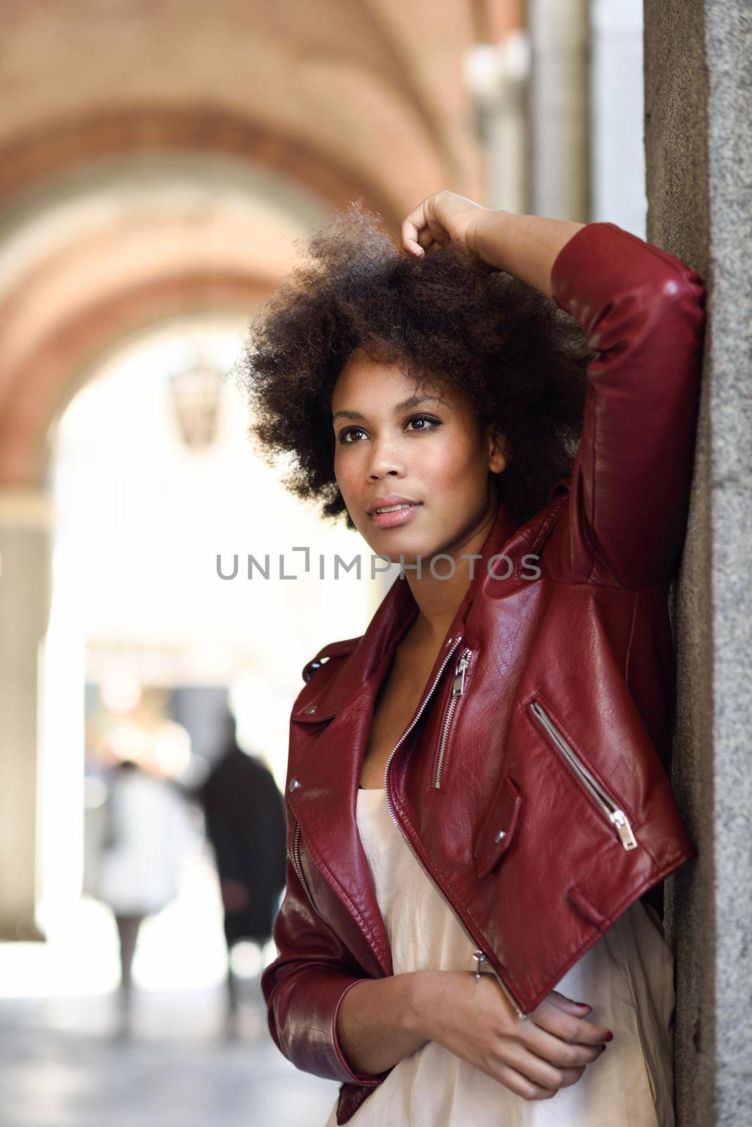 Young black female with afro hairstyle standing in an urban street. Mixed woman wearing red leather jacket and white dress with city background.