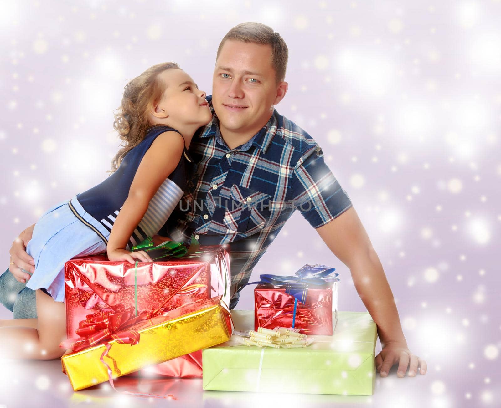 Gentle little girl kissing her beloved father, who is sitting on the floor surrounded by a variety of beautifully packaged boxes with gifts.Blue Christmas festive background with white snowflakes.