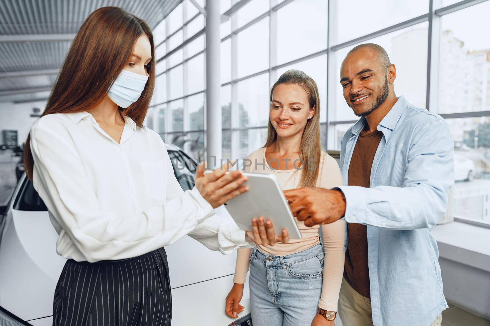 Car saleswoman wearing medical mask shows buyers couple something on digital tablet by Fabrikasimf