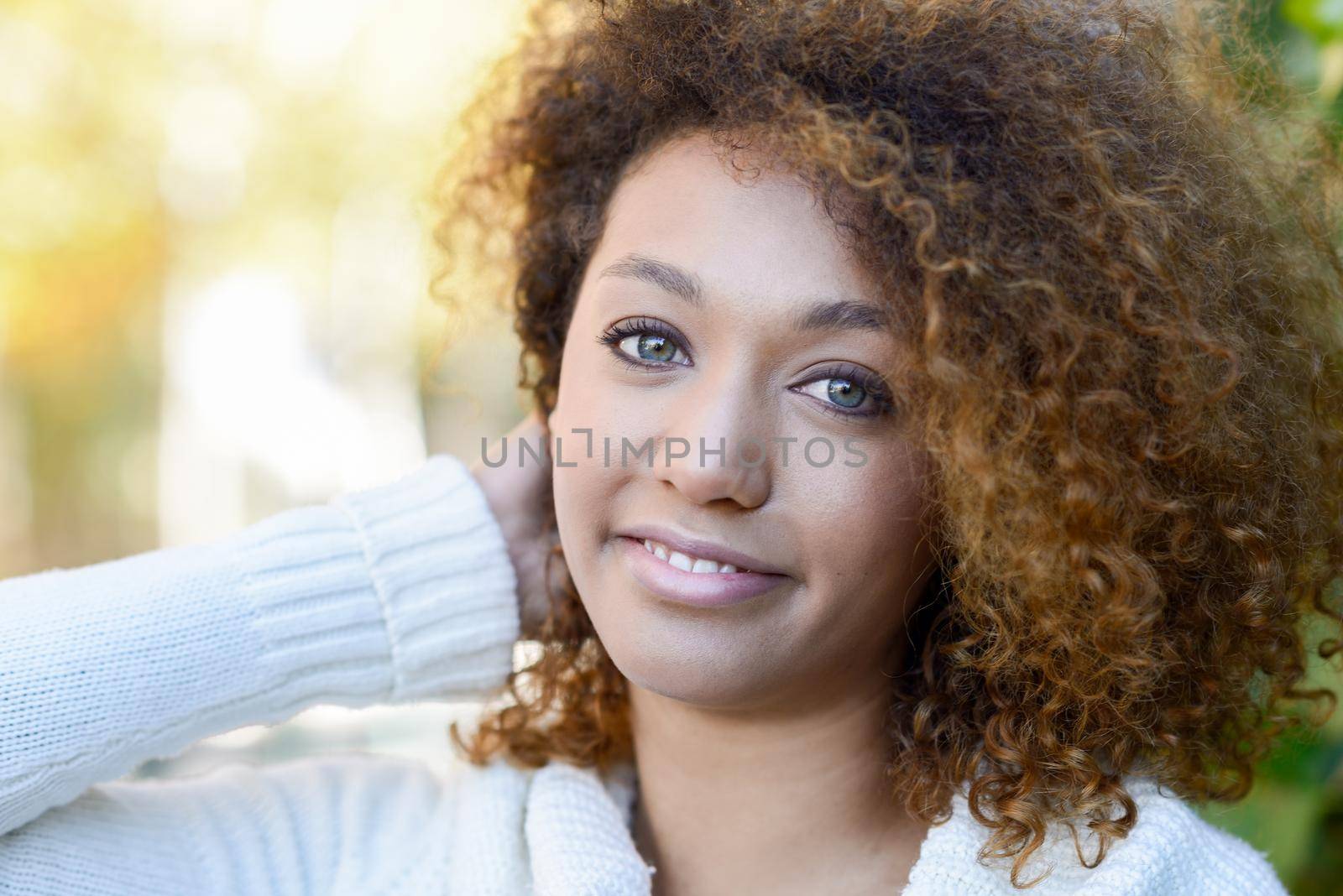 Beautiful young African American woman with afro hairstyle and green eyes wearing white winter dress with autumn leaves in the background.