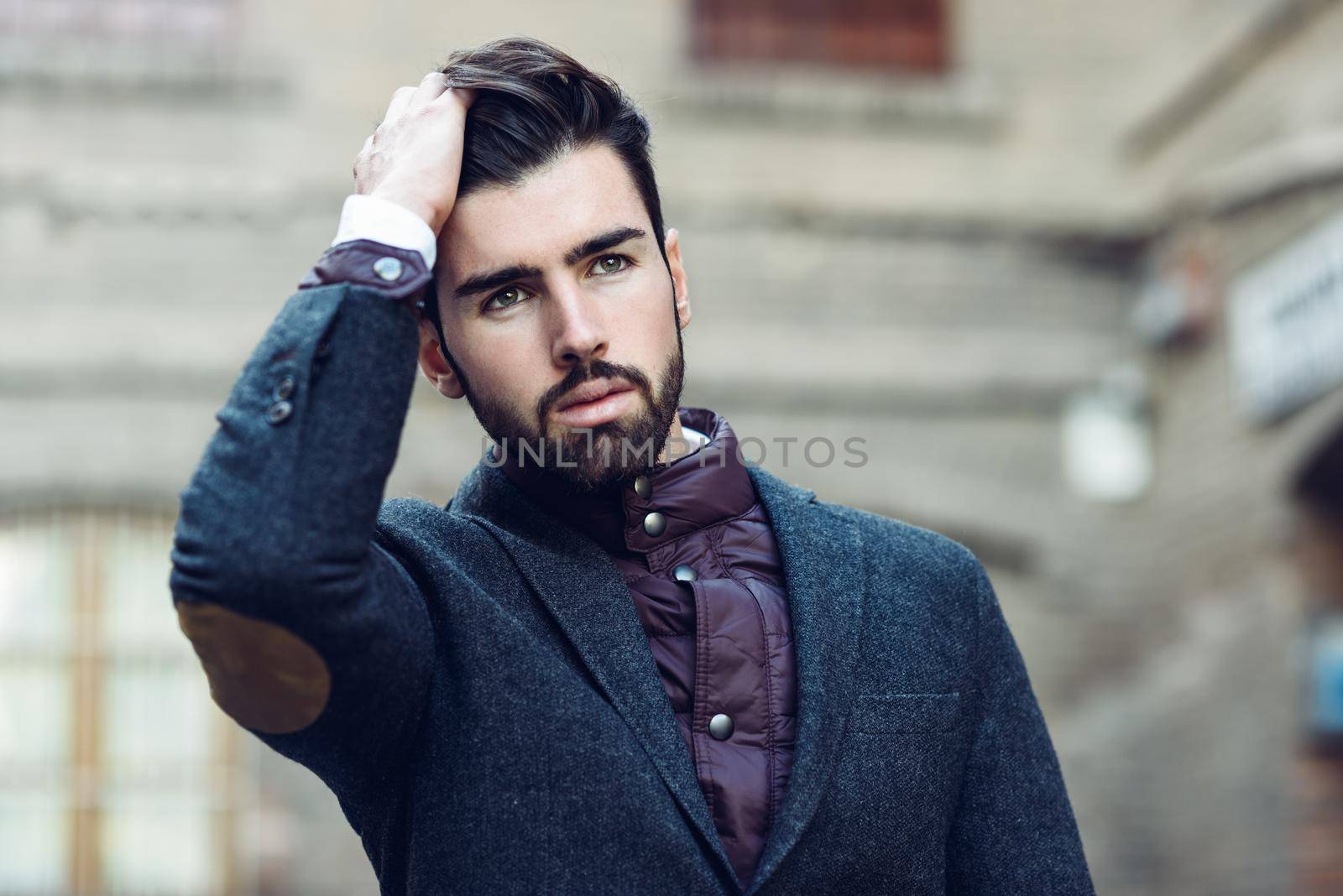 Young bearded man, model of fashion, in urban background wearing british elegant suit. Guy with beard and modern hairstyle touching his hair