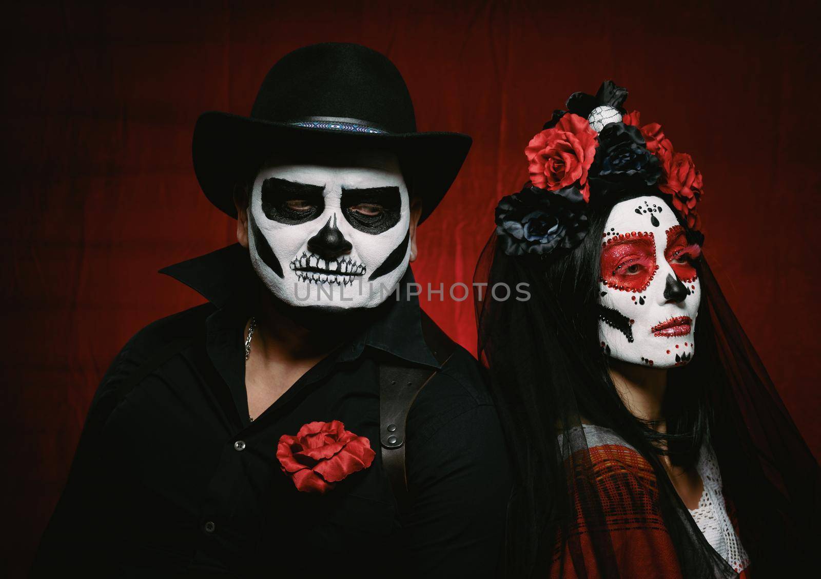 beautiful woman with a sugar skull makeup with a wreath of flowers on her head and a skeleton man in a black hat. Couple on dark red background