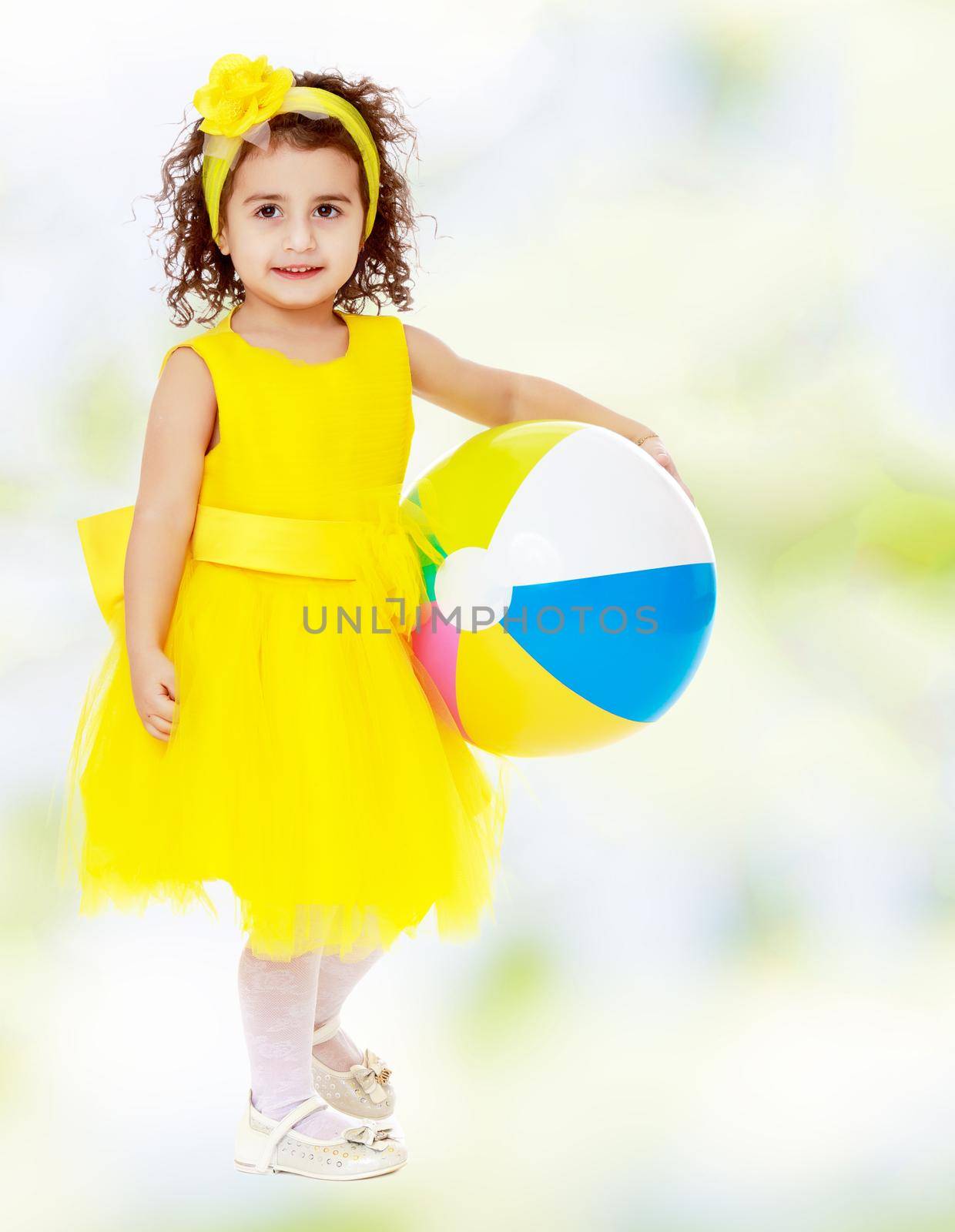 Cute curly girl in a bright yellow dress and bow on her head holding the ball. Close-up.white-green blurred abstract background with snowflakes.