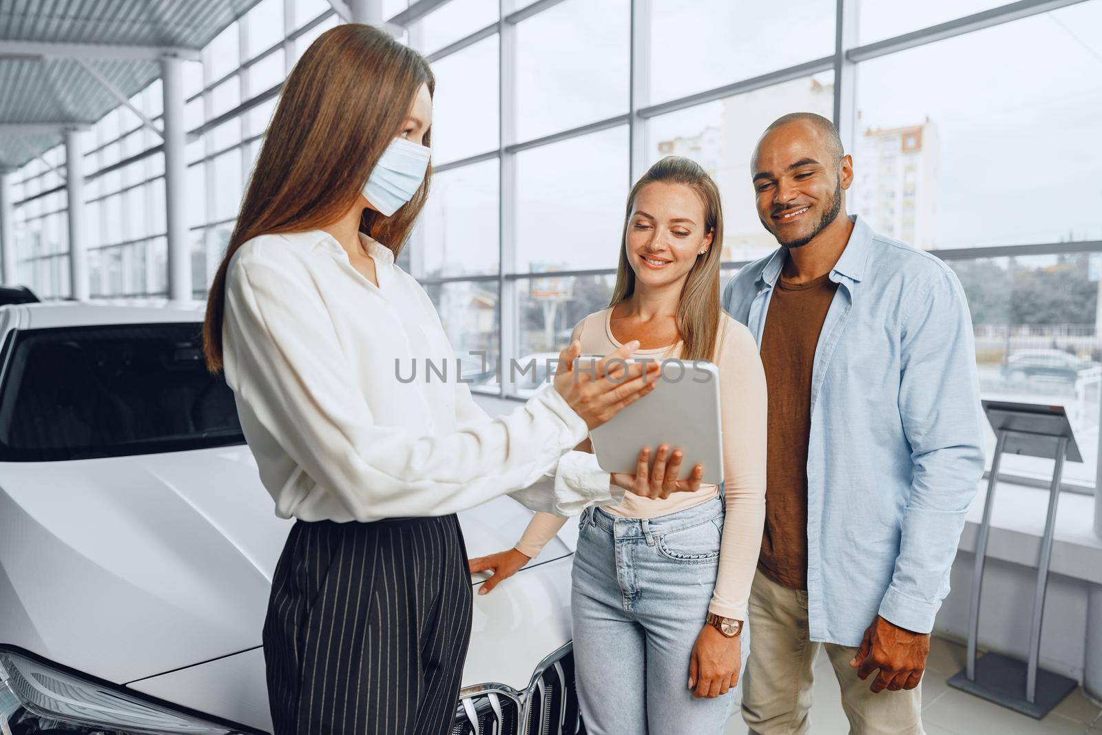 Car saleswoman wearing medical mask shows buyers couple something on digital tablet. .New pandemic job requirements concept