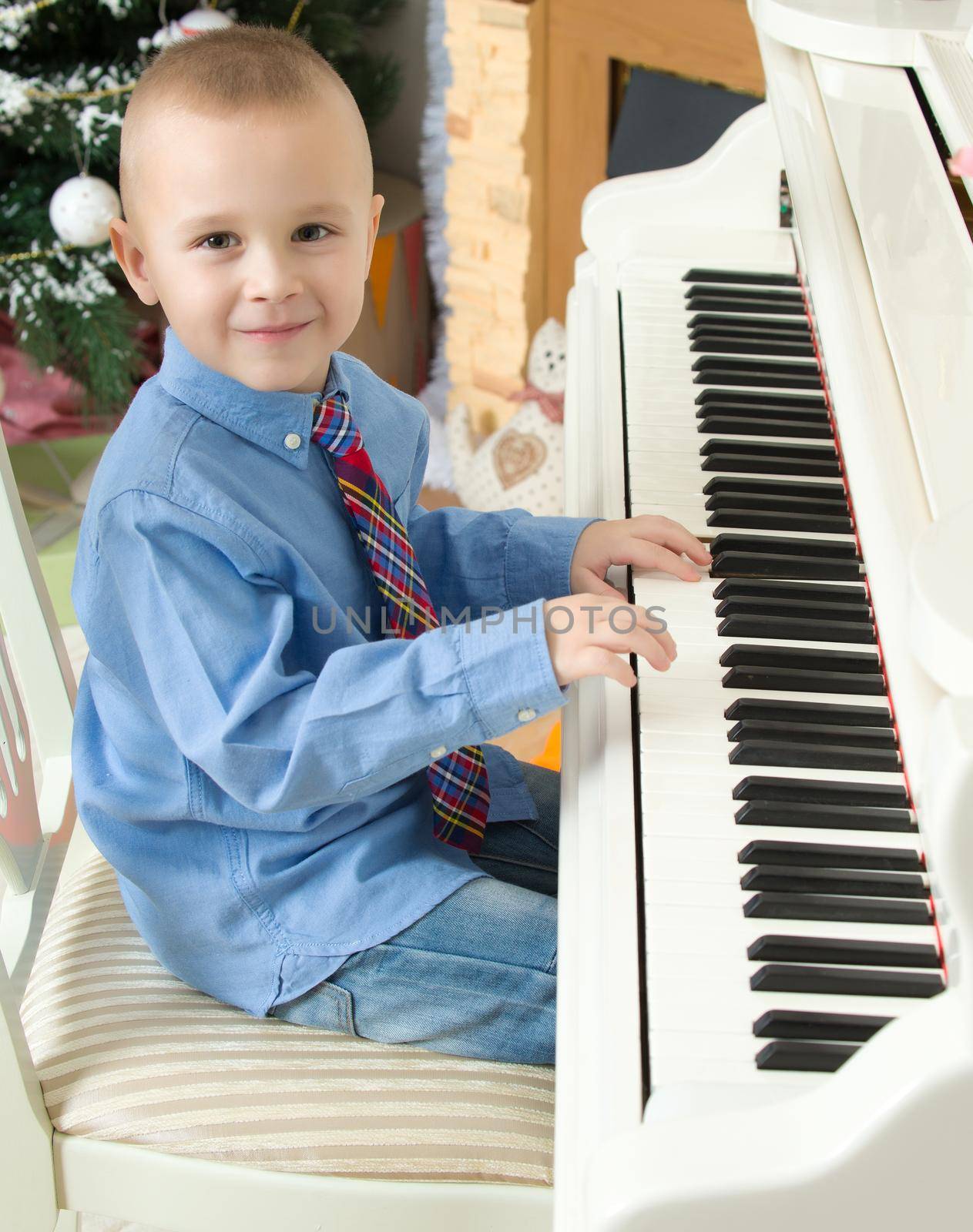 Elegant little boy in shirt and tie playing on a white Grand piano. Around the Christmas tree and the fireplace in the Christmas night.