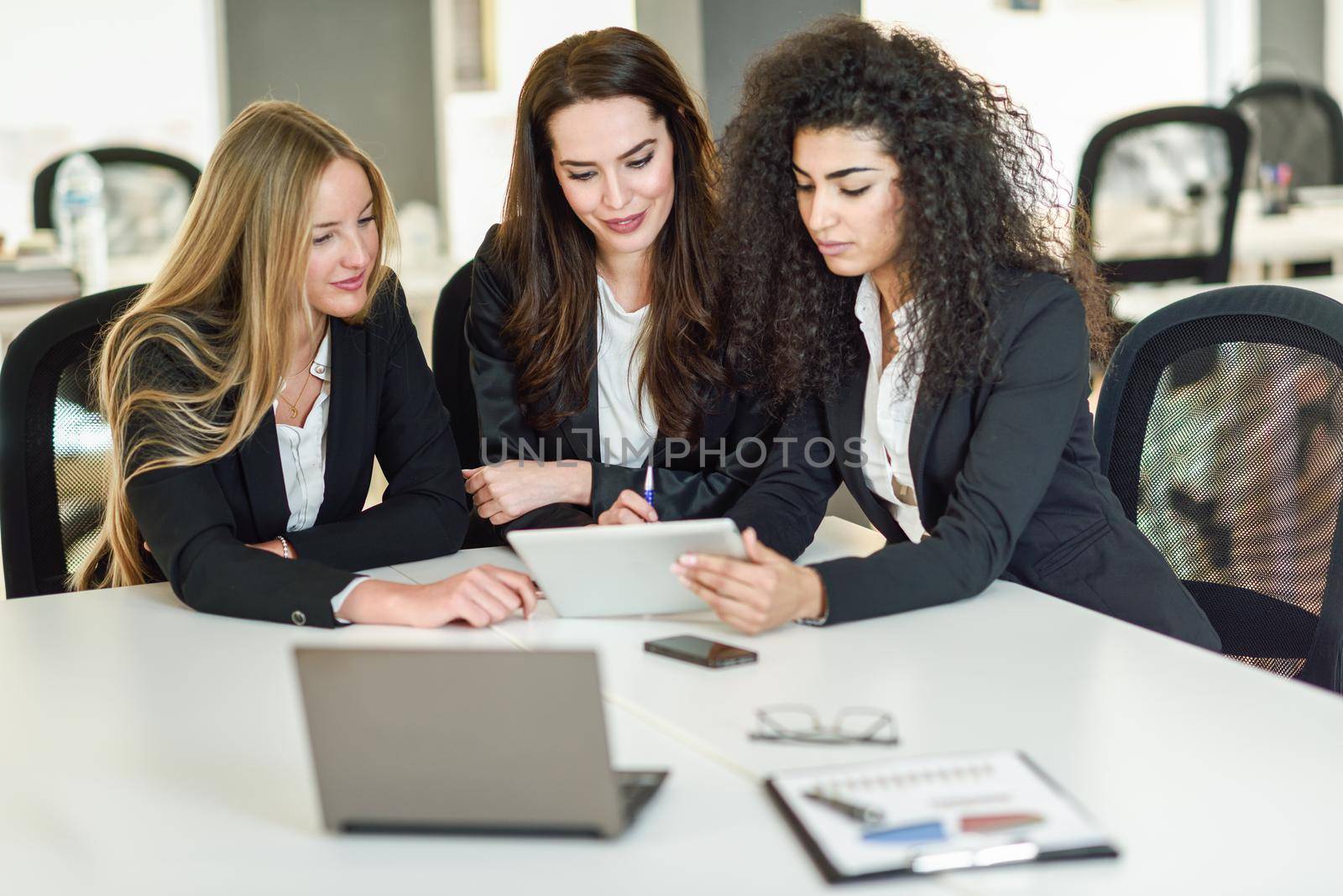 Three businesswomen working together in a modern office with white furniture. Teamwork concept. Caucasian, blonde, and muslim girls wearing suit. Multi-ethnic group of women