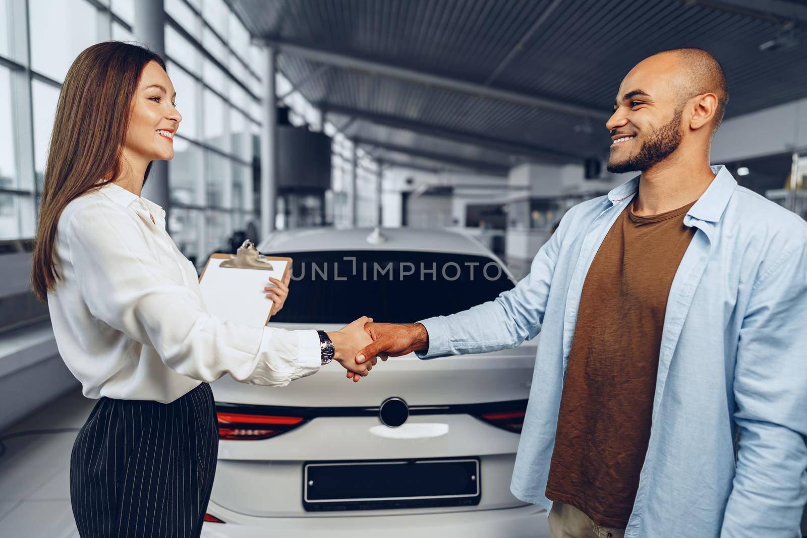 Woman car seller and man car buyer handshaking in car salon building