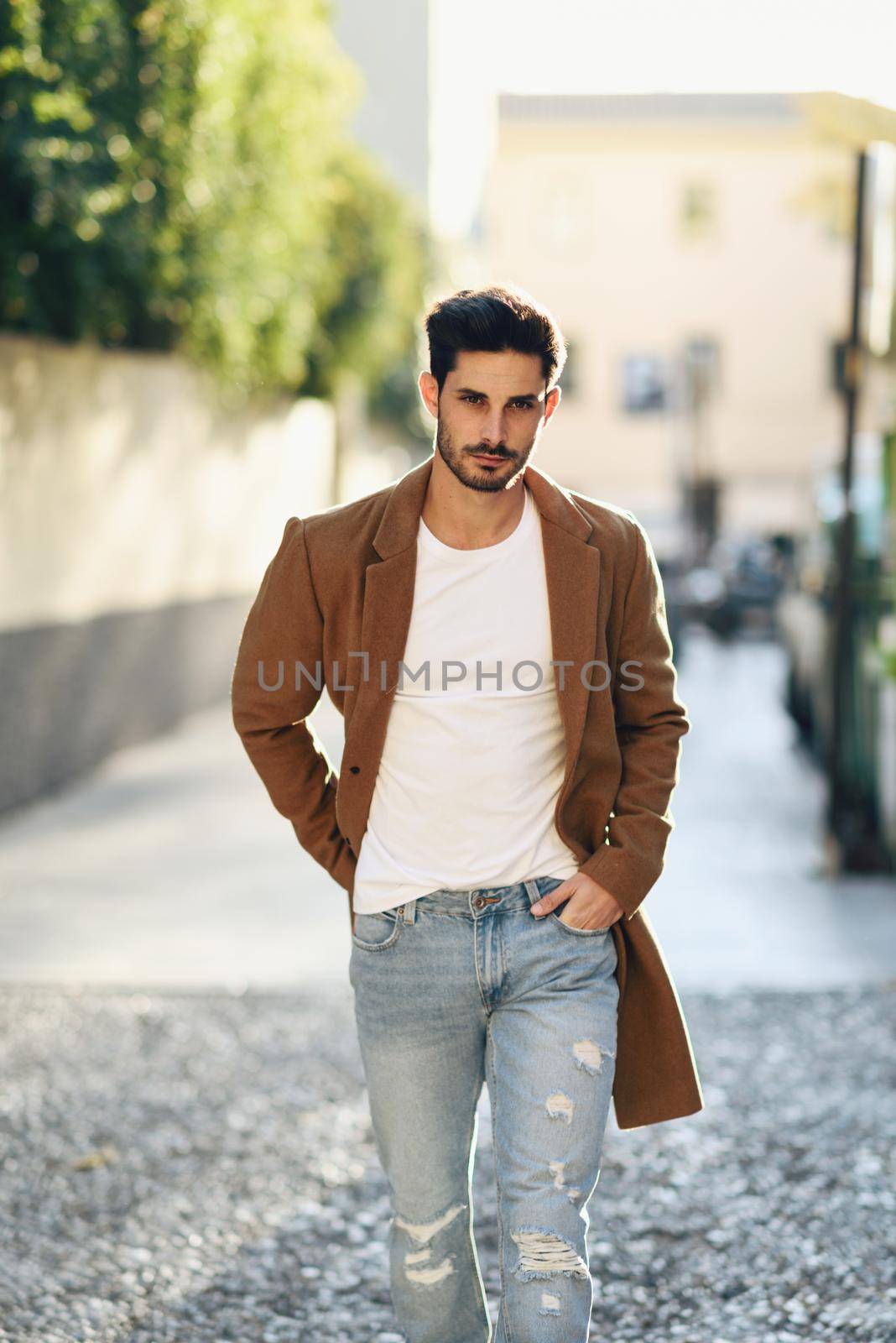 Young man wearing winter clothes in the street. Young bearded guy with modern hairstyle with coat, blue jeans and white t-shirt.