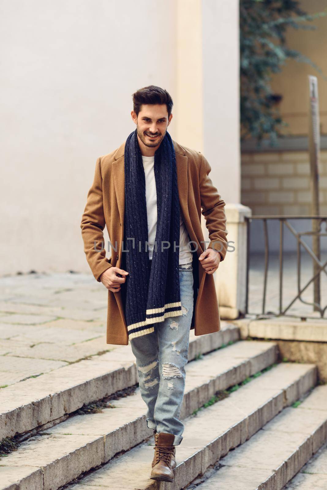 Young man wearing winter clothes in the street. Young bearded guy with modern hairstyle with coat, scarf, blue jeans and t-shirt.