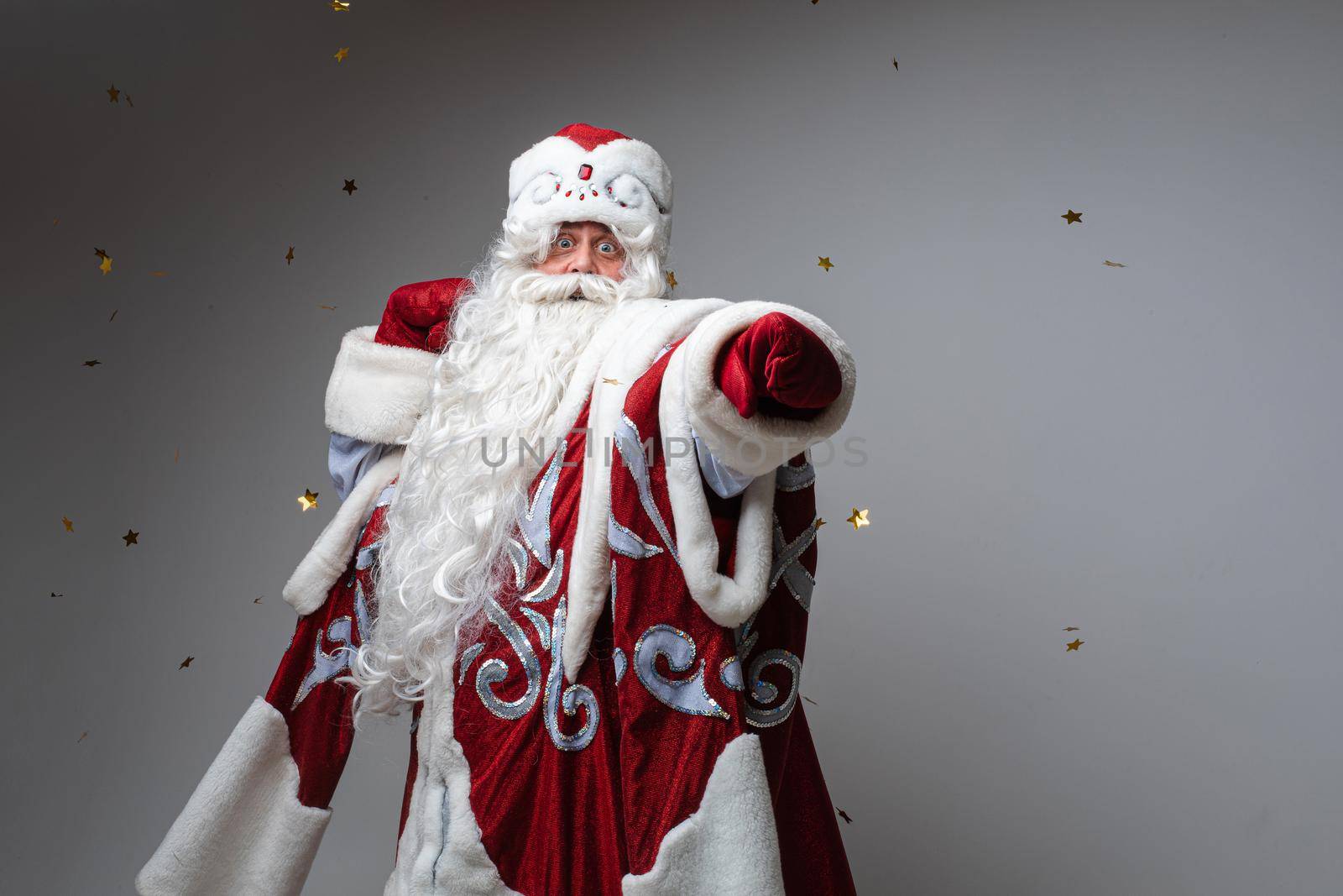 Funny senior male Santa Claus point by hand in red mitten to the camera on gray background with copy space. High quality photo