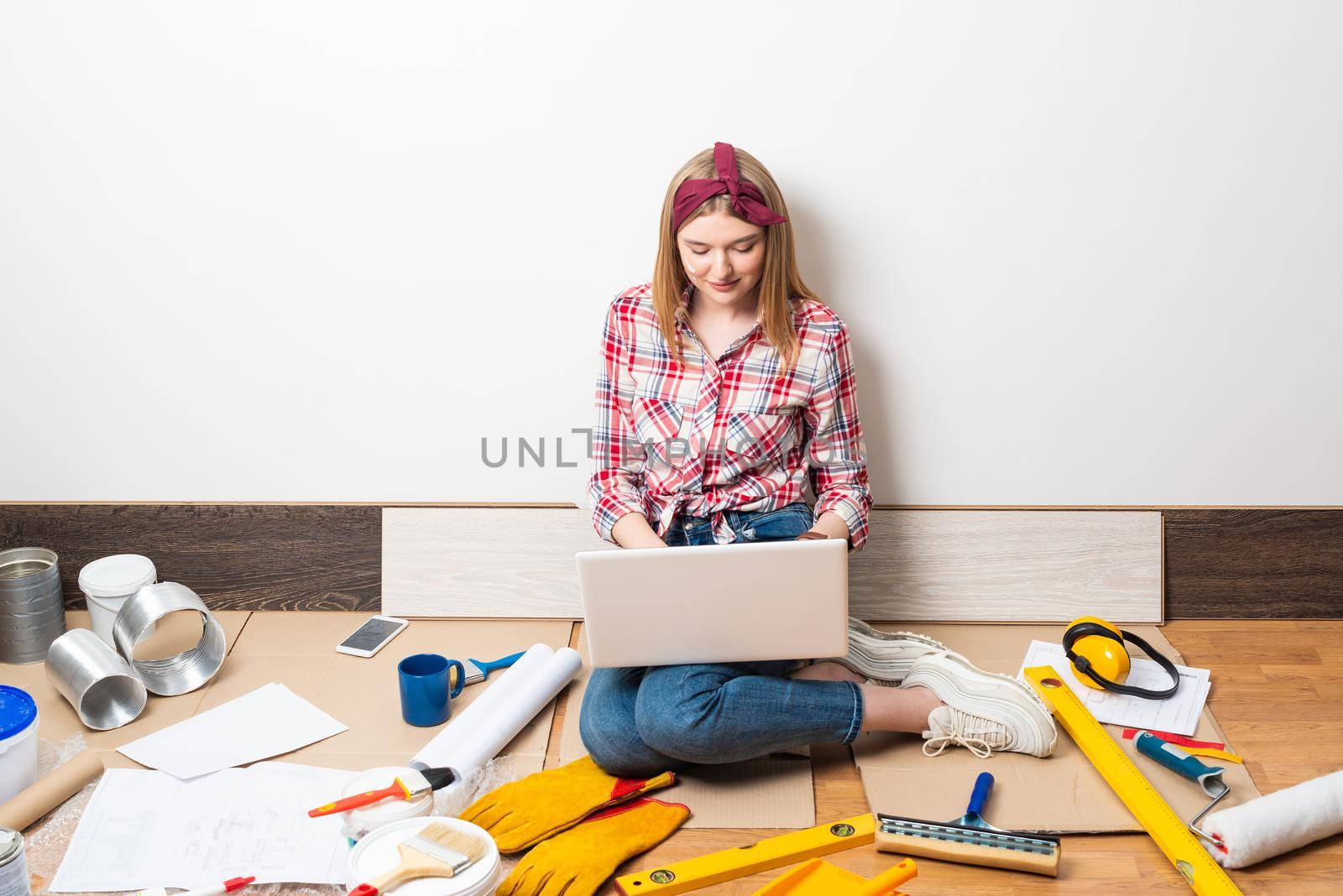 Beautiful girl using laptop at home. Apartment remodeling and house interior renovation concept with copy space. Young attractive woman in red checkered shirt and jeans sitting on floor with computer.