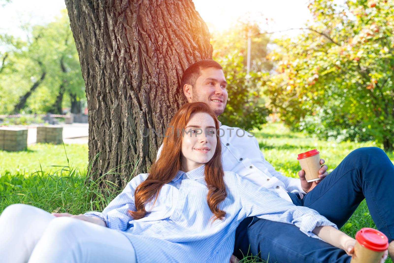 Young couple relaxing with coffee under tree in park on sunny day. Happy couple in love spend time outdoors together. Handsome man and pretty redhead girl sitting on green grass leaning against tree