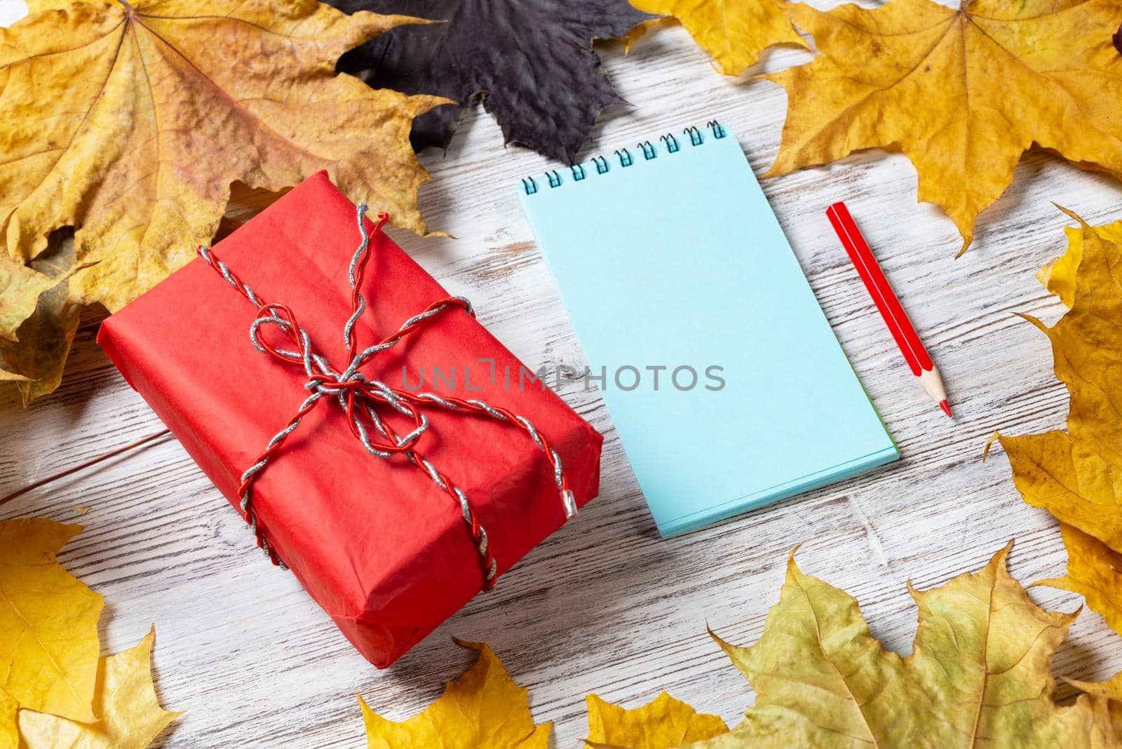 Spiral notepad with pen and gift box lies on vintage wooden desk with bright foliage. Flat lay composition with autumn leaves on white wooden surface. Happy thanksgiving holiday congratulation.