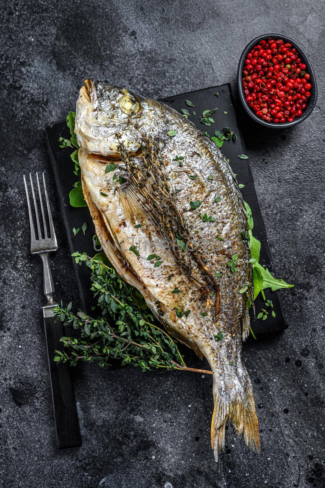 Roasted sea bream fish with herbs on a cutting board. Black background. Top view.