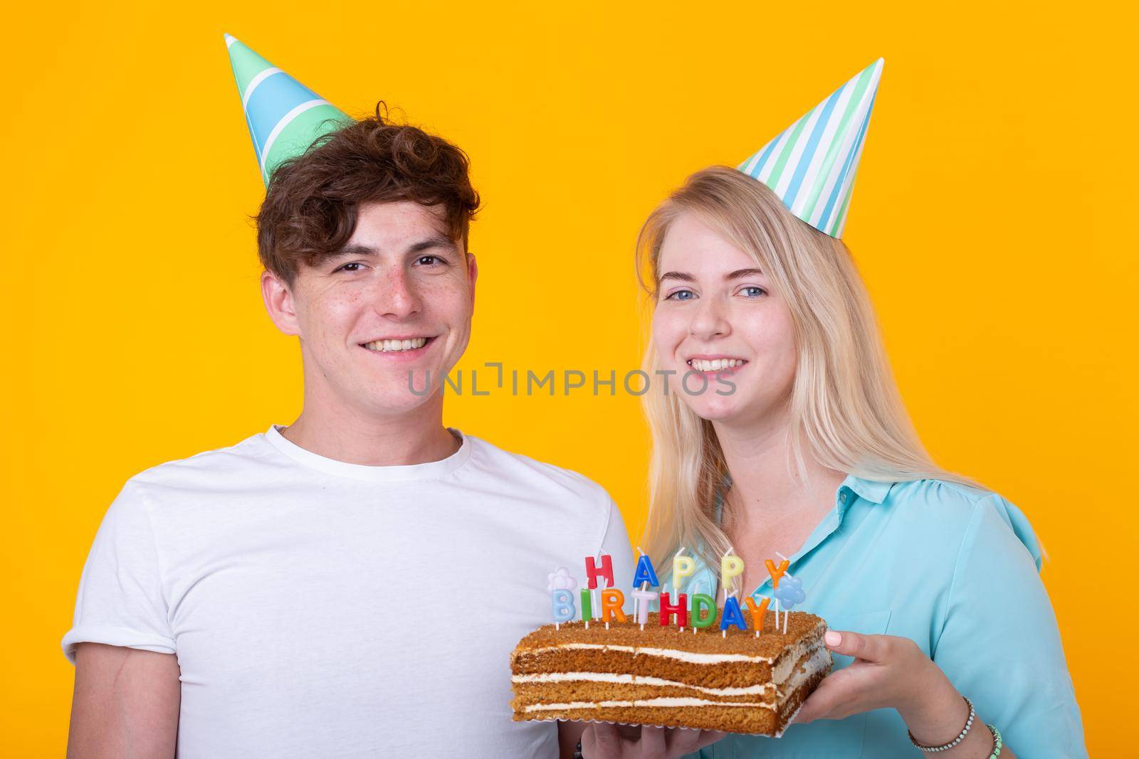 Funny young couple in paper caps and with a cake make a foolish face and wish happy birthday while standing against a yellow background. Concept of congratulations and fooling around