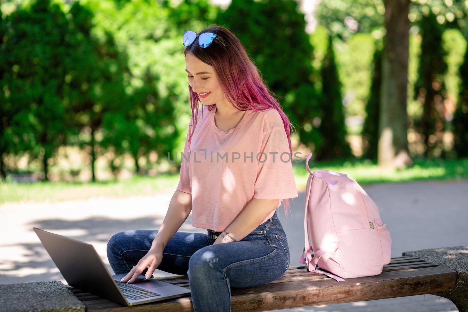 Young girl with pink hair is studying in the spring park, sitting on the wooden bench and browsing on her laptop