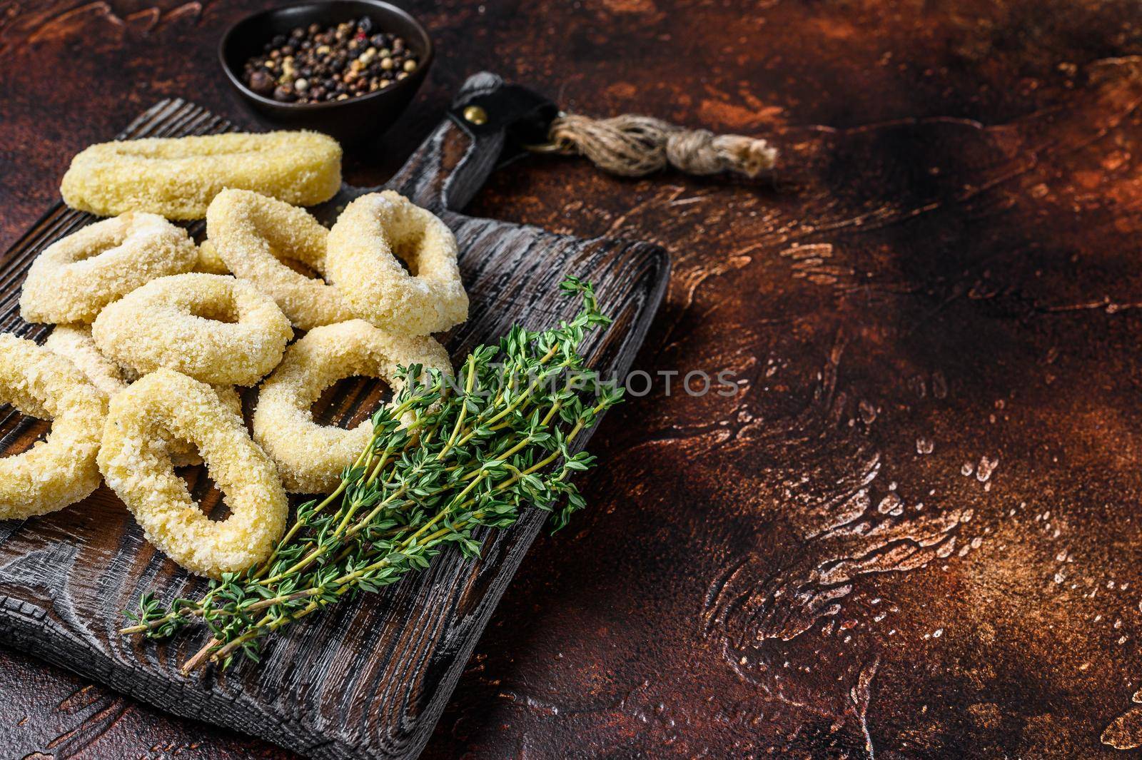 Frozen raw squid rings in breadcrumbs on a cutting board. Dark background. Top view. Copy space by Composter