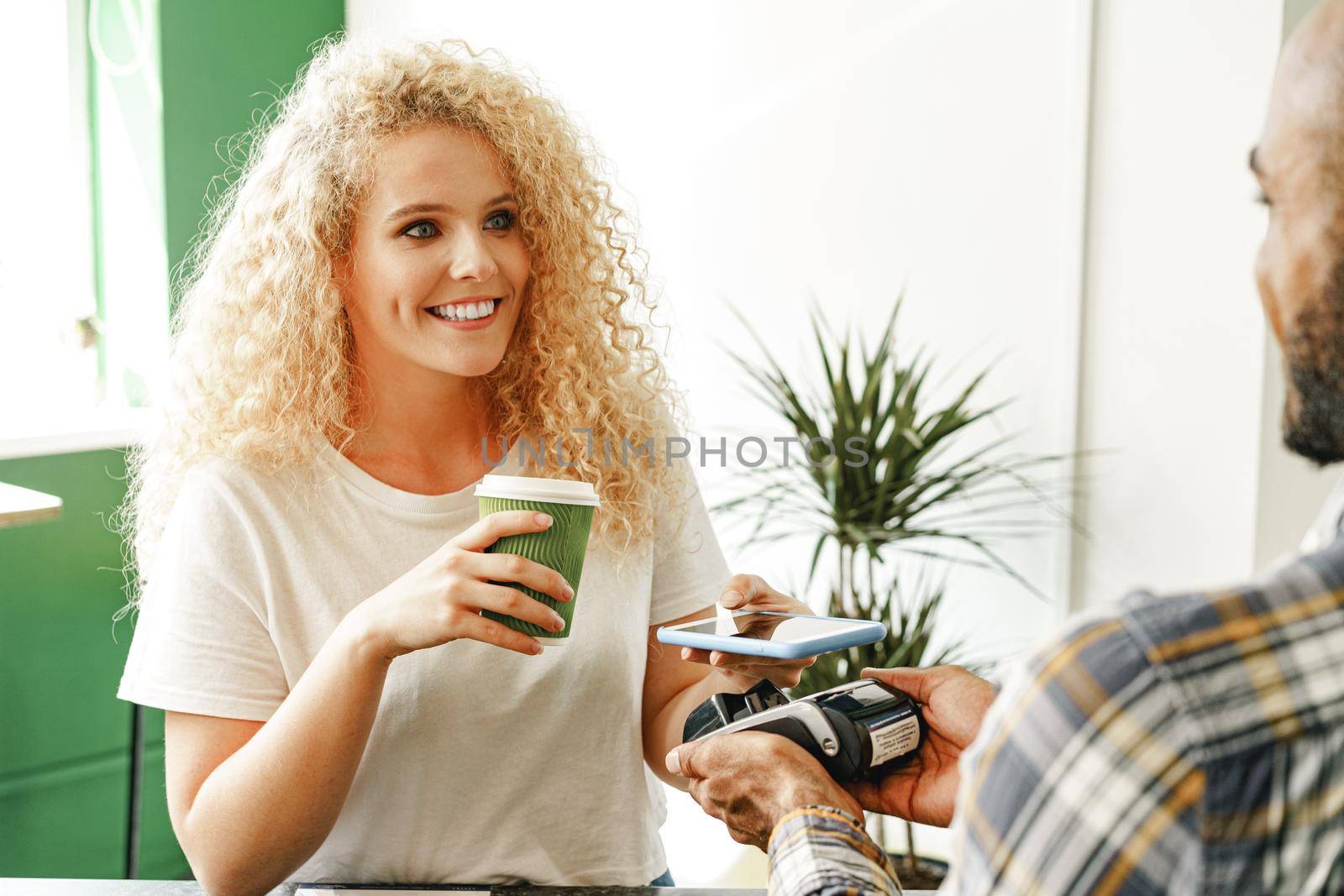 Woman customer of coffee shop paying for coffee through mobile phone using contactless technology by Fabrikasimf