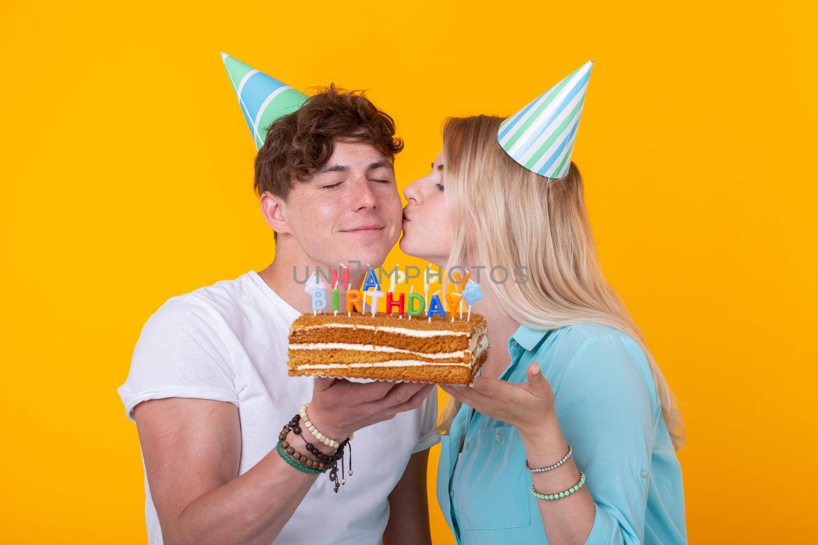 Funny young couple in paper caps and with a cake make a foolish face and wish happy birthday while standing against a yellow background. Concept of congratulations and fooling around