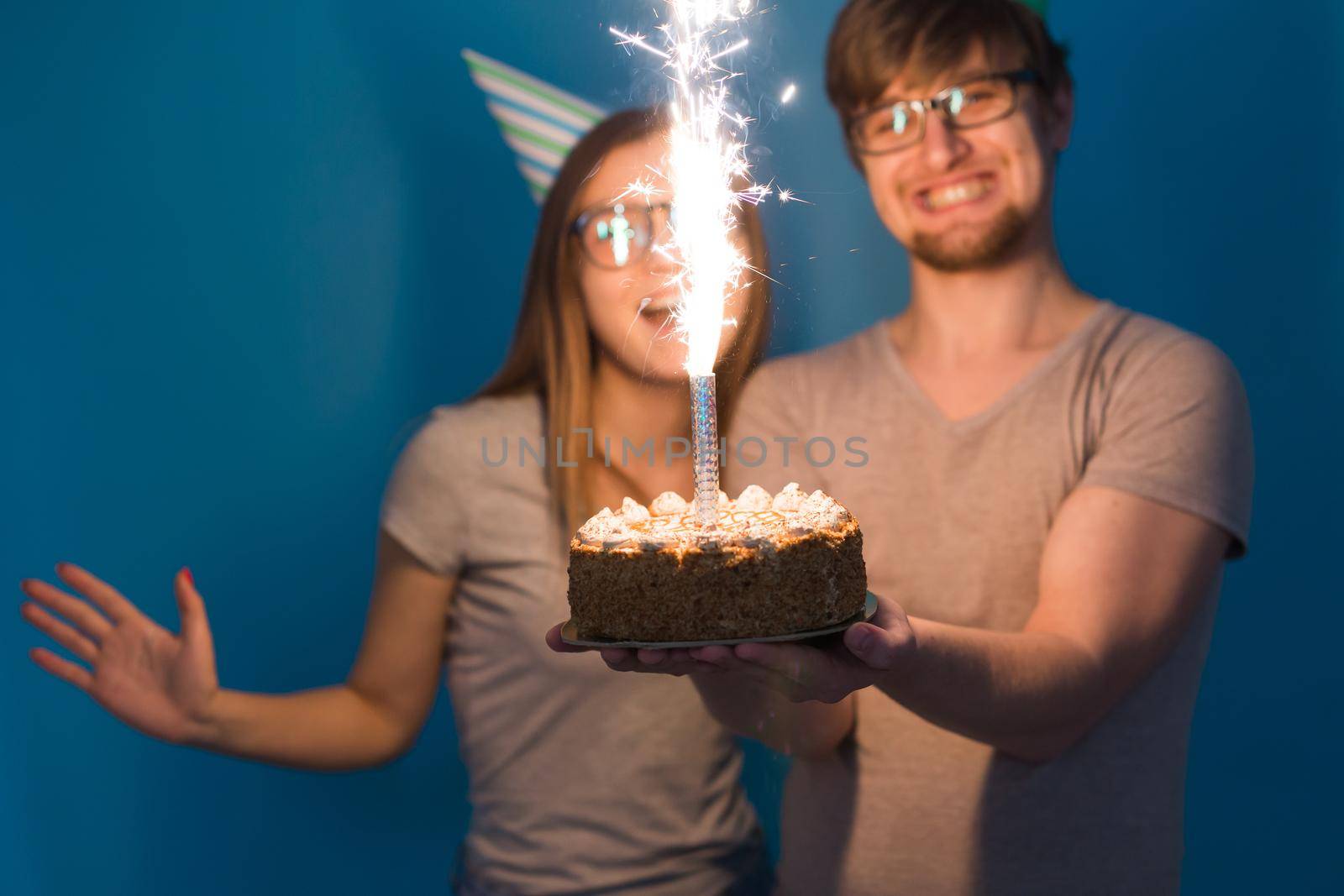 Funny young couple in paper caps and with a cake make a foolish face and wish happy birthday while standing against a blue background. Concept of congratulations and fooling around