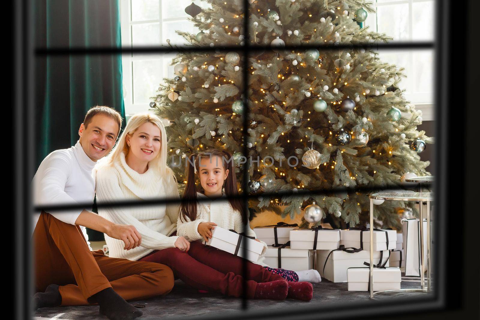 Young big family celebrating Christmas enjoying dinner, view from outside through a window into a decorated living room with tree and candle lights, happy parents eating with three kids