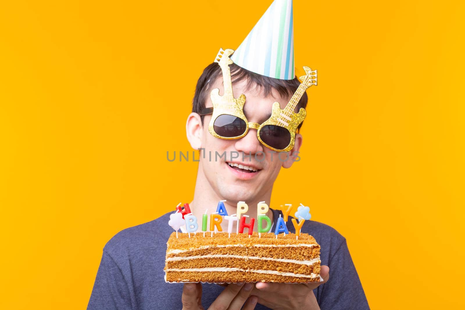 Portrait of a funny positive guy with a paper cap and glasses holding a congratulatory cake in his hands on a yellow background. Concept and fun and celebration. Advertising space