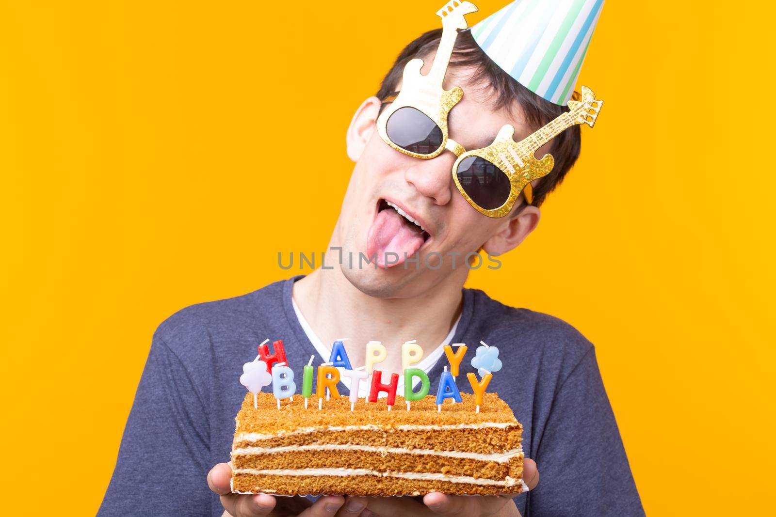 Positive funny young asian guy with a cap and a burning candle and a cake in his hands posing on a yellow background. Anniversary and birthday concept. Advertising space