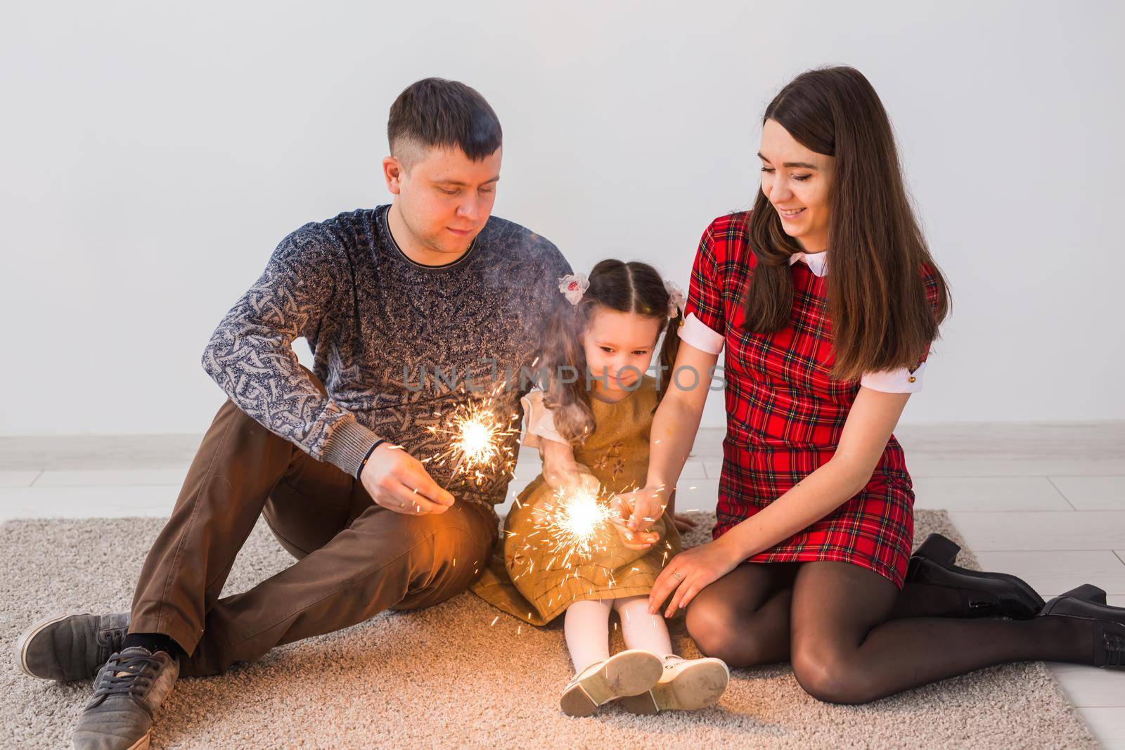 Celebration, family and holidays concept - happy parents and little daughter with sparklers sitting on carpet over grey background.