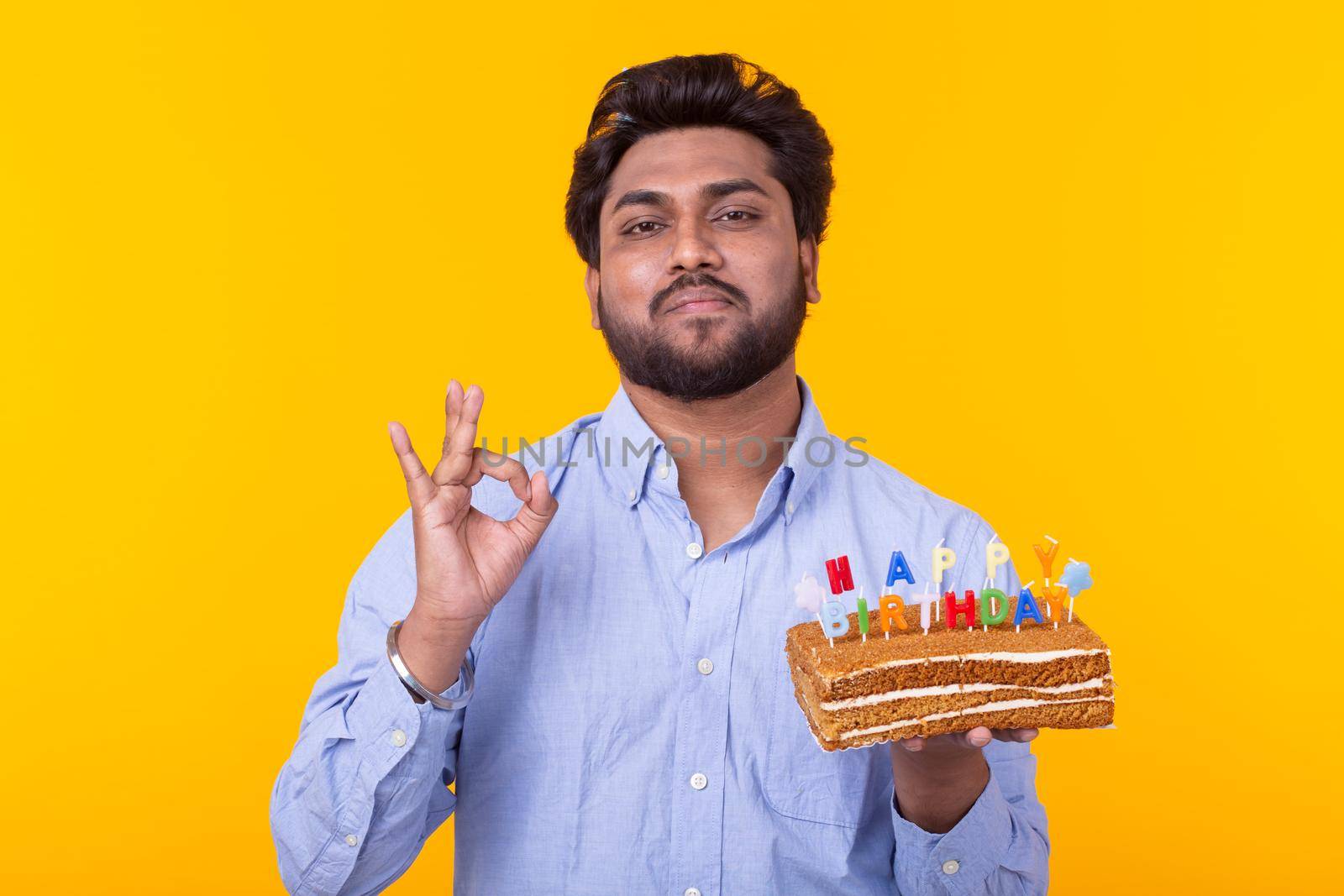 Positive young man holding a happy birthday cake and two burning bengal lights posing on a yellow background. Advertising space