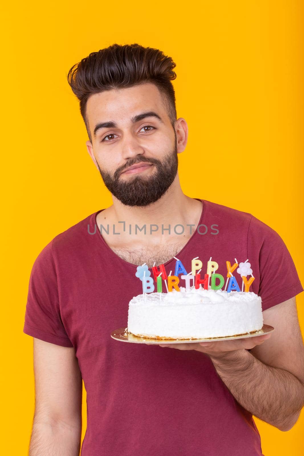 Embarrassed young male hipster with beard holding a birthday cake in his hands and looking thoughtfully at him posing on a yellow background. Concept of time quickly flies by. Advertising space