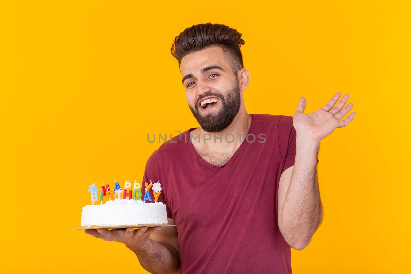 Positive handsome young hipster man in burgundy t-shirt holding congratulatory cake with inscription happy birthday posing on a yellow background. Concept of congratulations and anniversaries