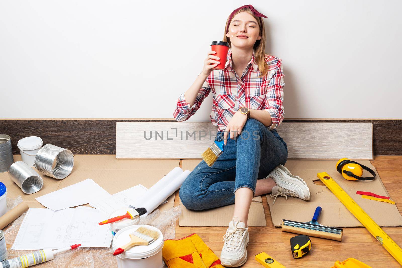 Dreamy girl painter relaxing on floor with cup of coffee. House remodeling and interior renovation concept with copy space. Young woman in red checkered shirt and jeans planning to redesign her home.