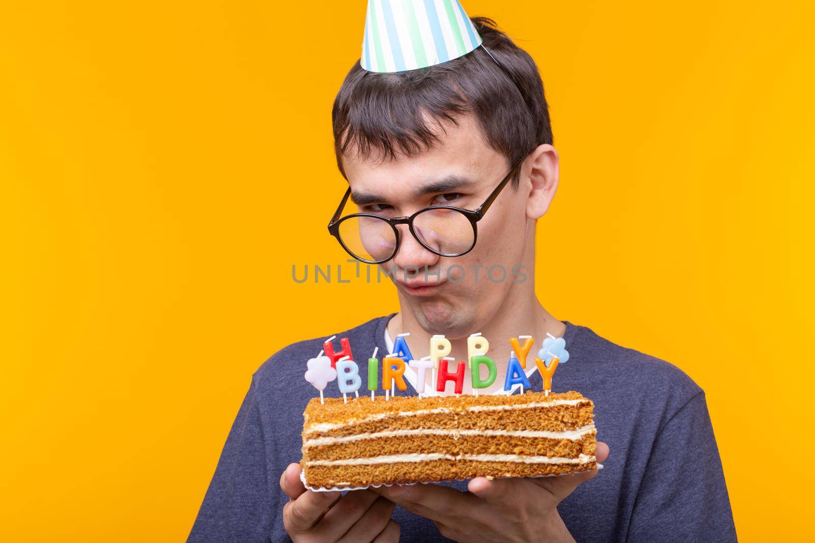 Positive funny young asian guy with a cap and a burning candle and a cake in his hands posing on a yellow background. Anniversary and birthday concept. Advertising space