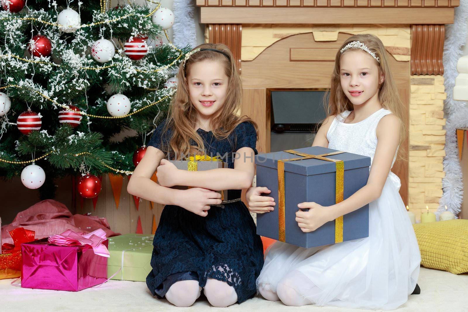 Cute little twin girls, sitting on the floor near the Christmas tree and electric fireplace on which candles are burning. In the hands of the girls boxes with gifts.