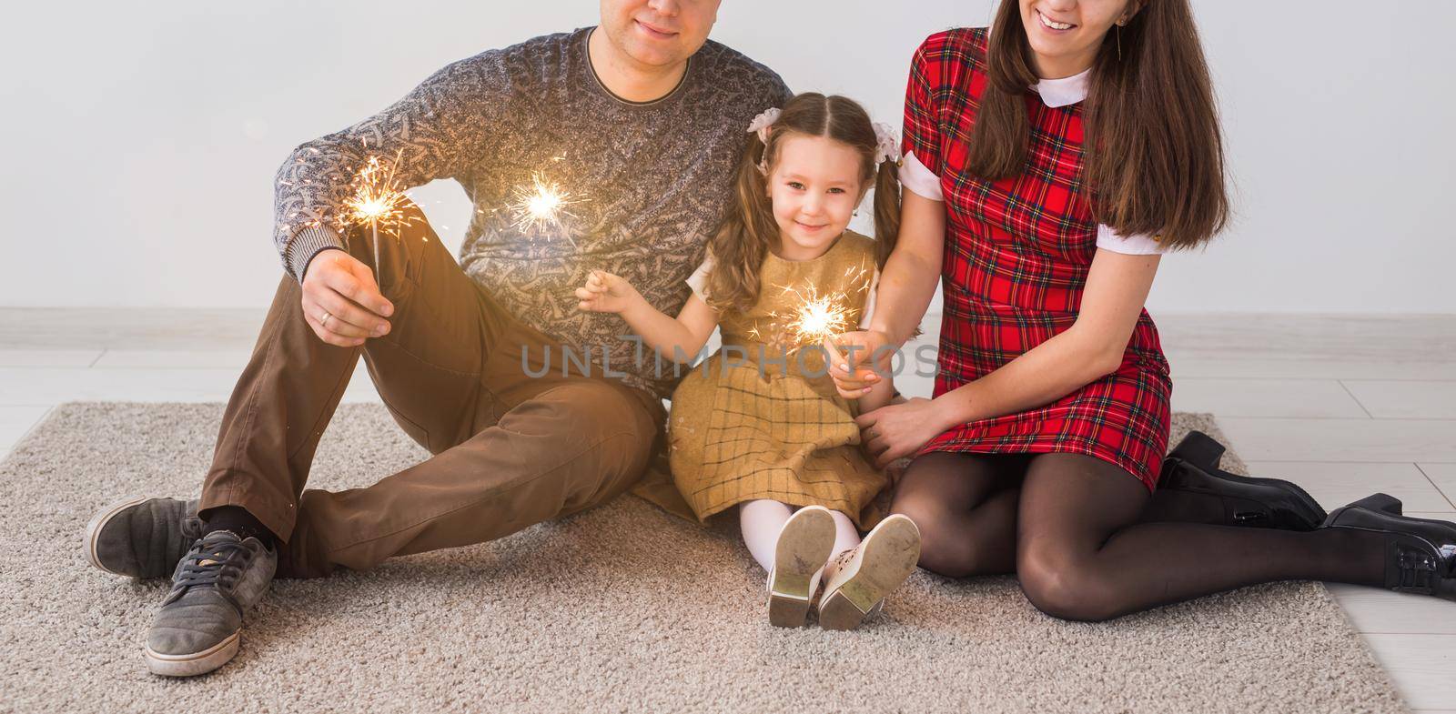 Celebration, family and holidays concept - happy parents and little daughter with sparklers sitting on carpet over grey background.