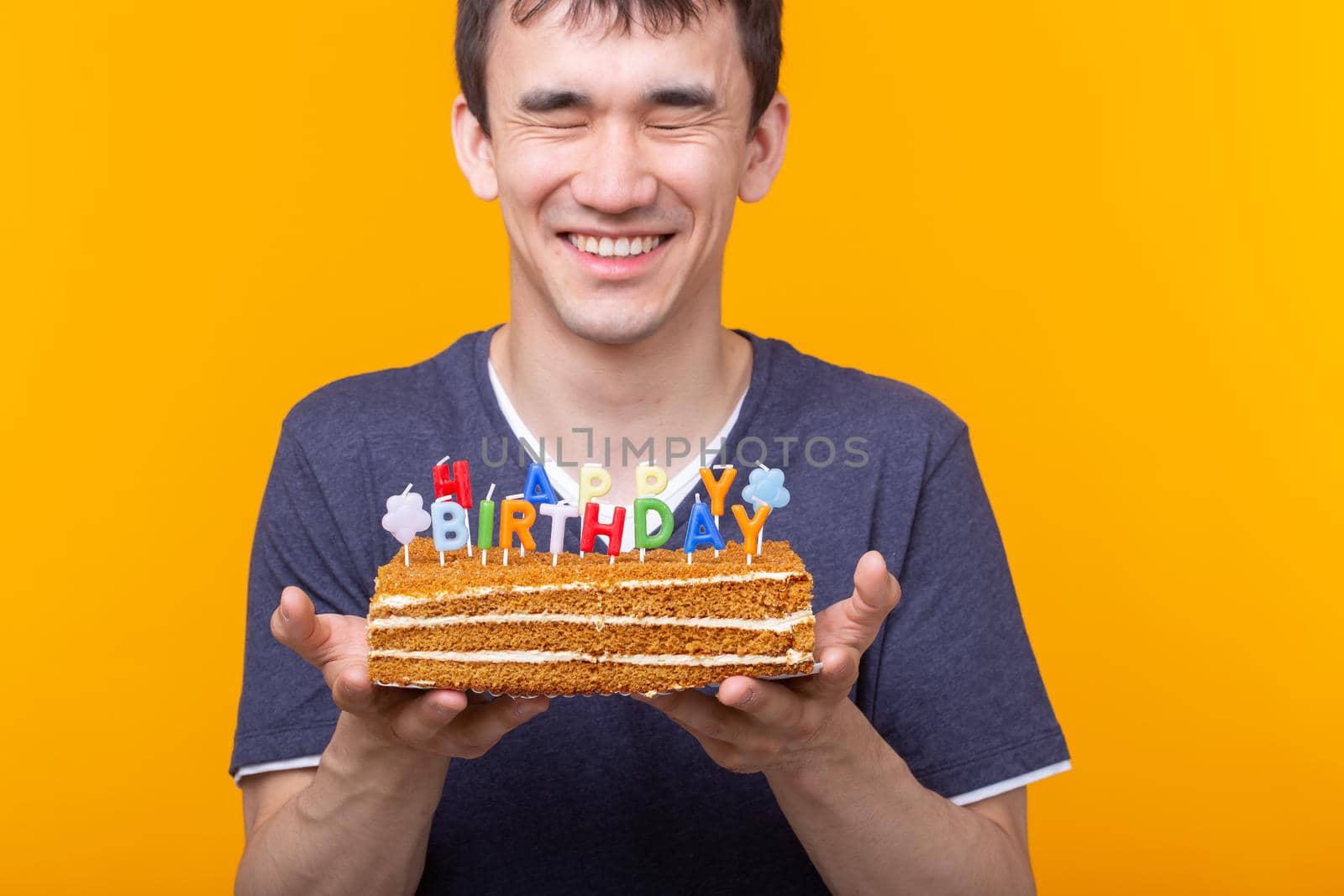 Crazy cheerful young guy with glasses holding a burning candle in his hands and a congratulatory cake on a yellow background. Birthday and anniversary celebration concept. Advertising space