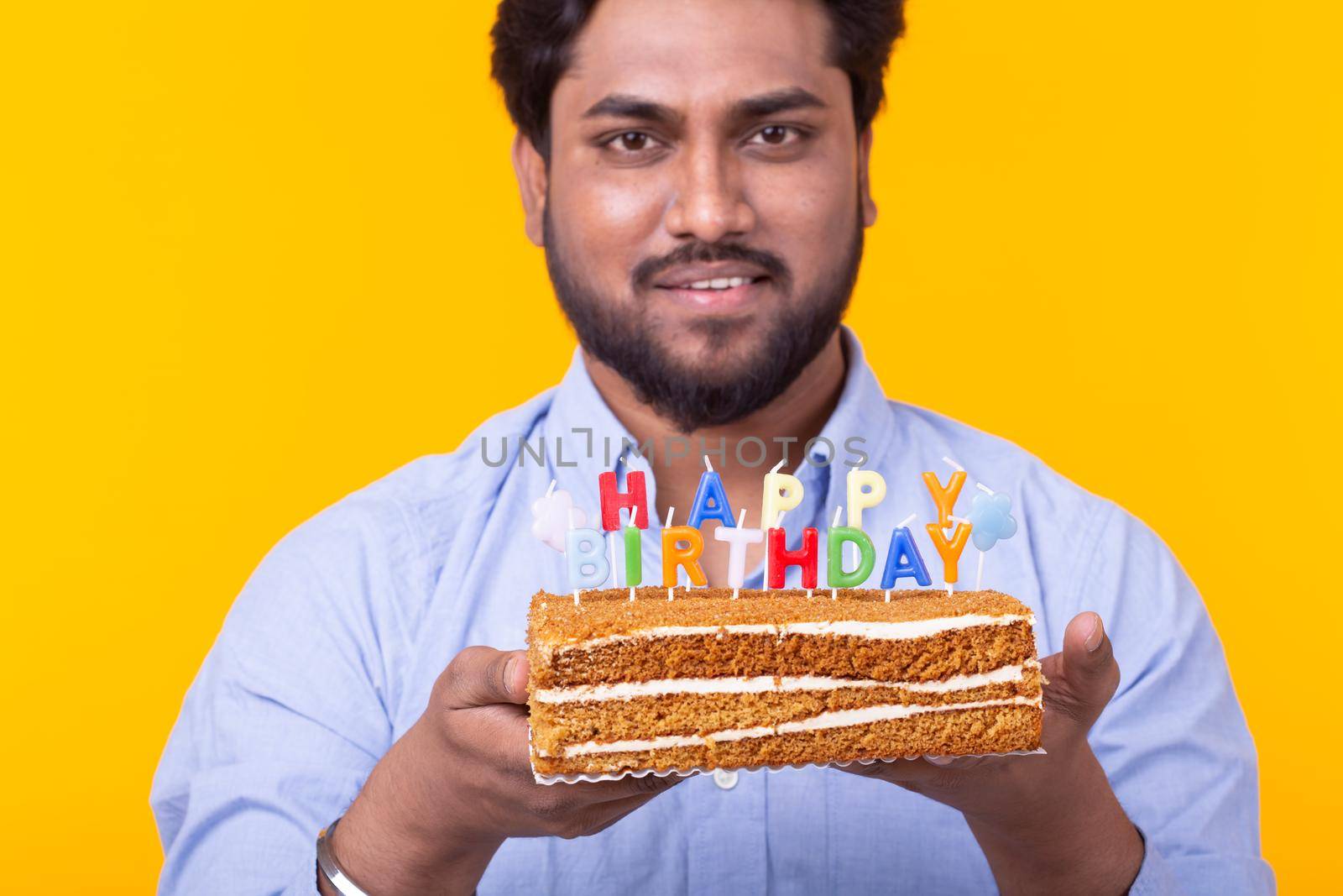 Positive young man holding a happy birthday cake and two burning bengal lights posing on a yellow background. Advertising space