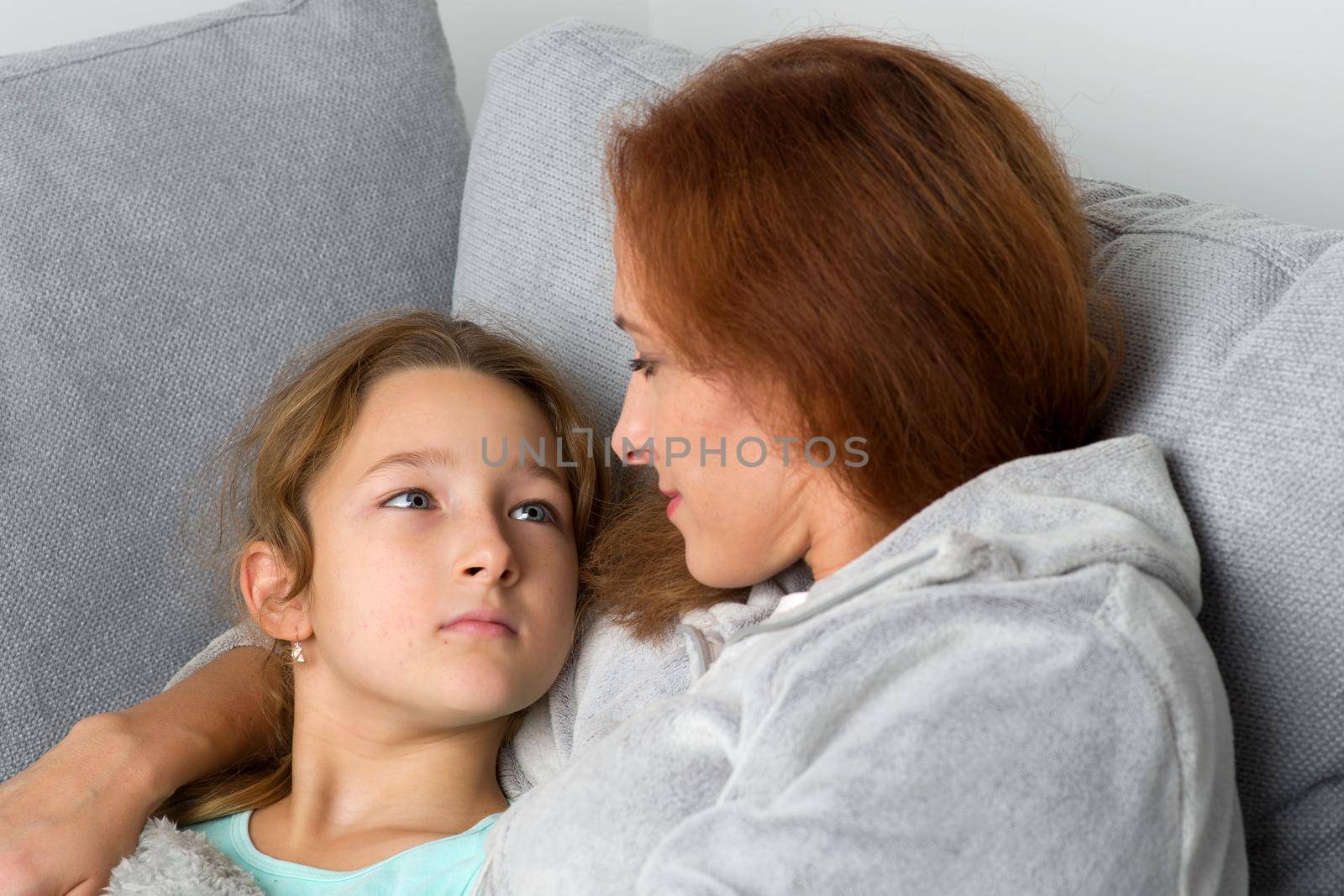 Mom and her daughter looking at each other. Loving mother hugging her preteen daughter. Cheerful woman and child wearing indoor clothes sitting together on couch at home