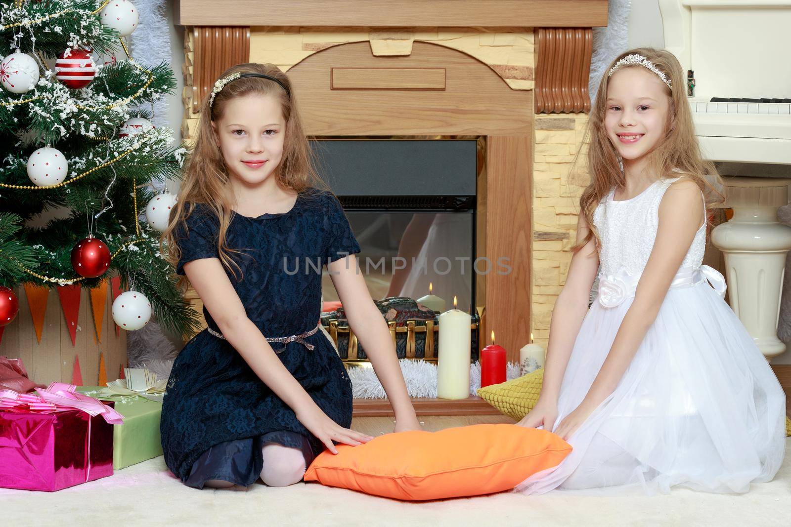 Cute little twin girls, sitting on the floor near the Christmas tree and electric fireplace on which candles are burning.