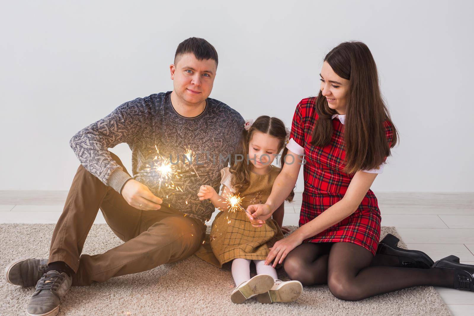 Celebration, family and holidays concept - happy parents and little daughter with sparklers sitting on carpet over grey background.