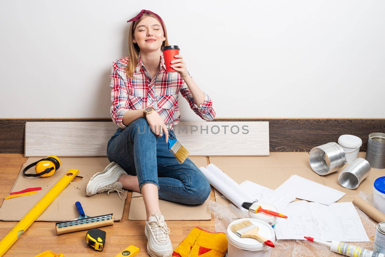 Dreamy girl painter relaxing on floor with cup of coffee. House remodeling and interior renovation concept with copy space. Young woman in red checkered shirt and jeans planning to redesign her home.