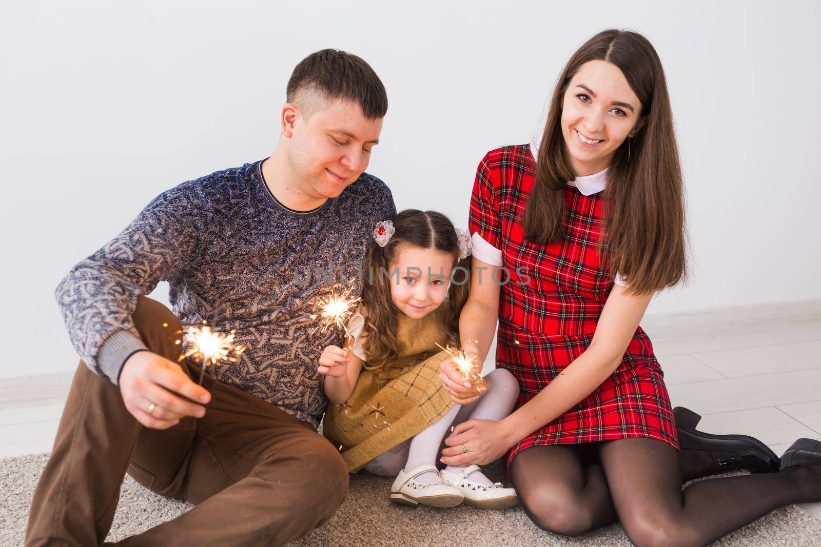 Celebration, family, new year, christmas and holidays concept - happy parents and little daughter with sparklers sitting on carpet over grey background by Satura86