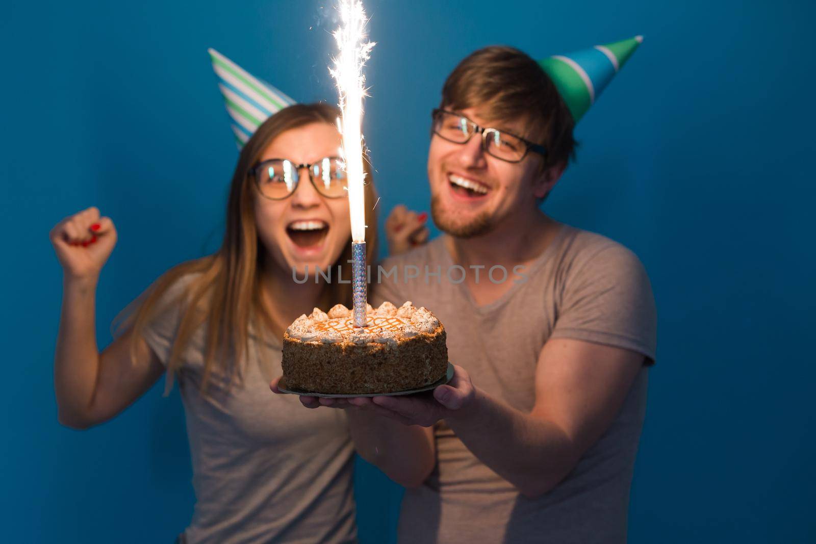 Funny young couple in paper caps and with a cake make a foolish face and wish happy birthday while standing against a blue background. Concept of congratulations and fooling around