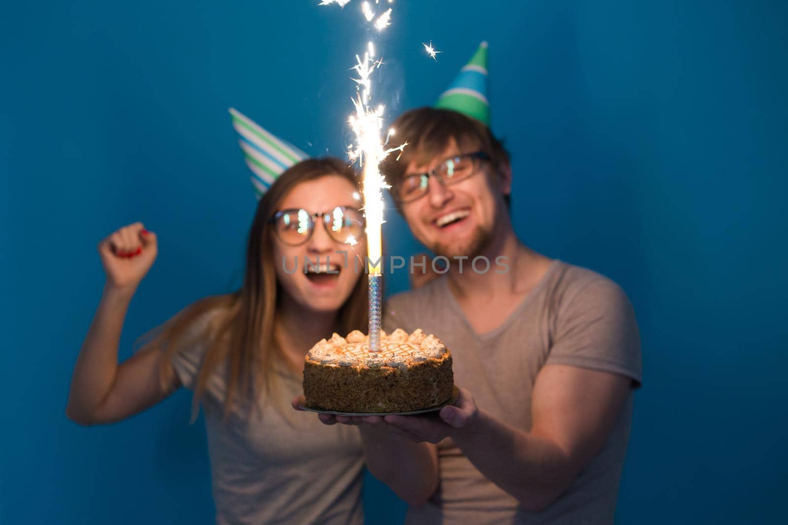 Young cheerful students charming girl and nice guy in greeting paper caps holding a cake with a bengal sparks candle. Concept of congratulations on the holiday and anniversary